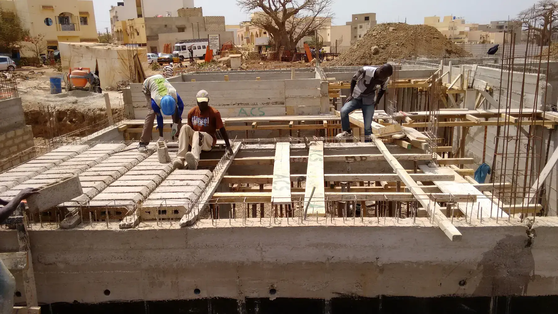 Slab formwork with hollow-block infill and rebar being laid by workers, basement walls below