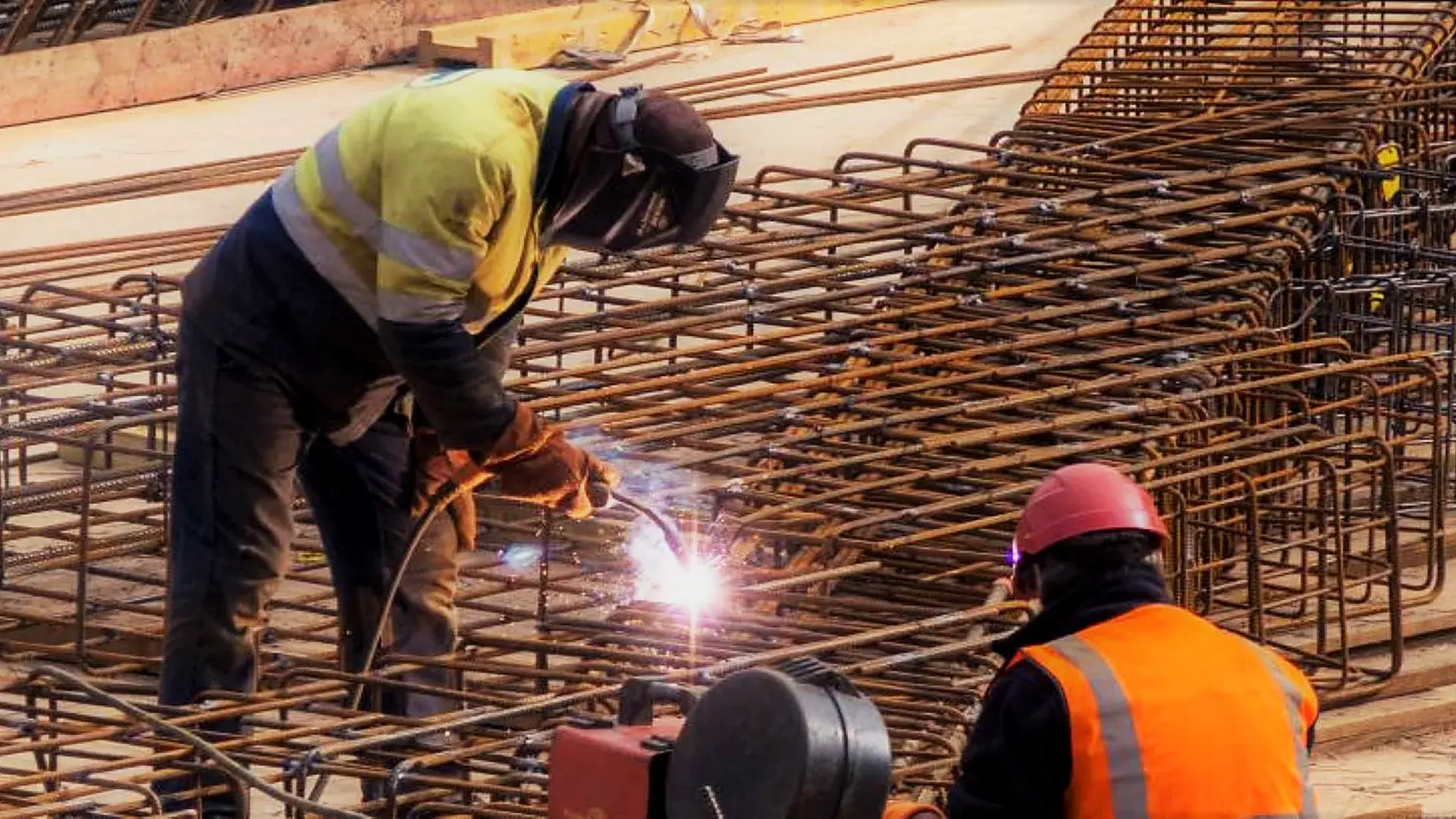 Soudeur assemblant des cages d'armatures sur un chantier béton, avec un ouvrier au premier plan